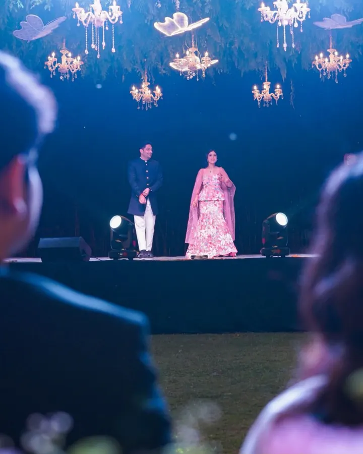 A couple on a grand stage under hanging chandeliers, with guests watching